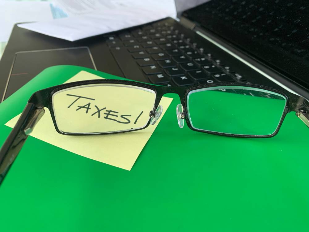 A pair of glasses rests on a green surface with a sticky note labeled "TAXES" beside a laptop keyboard, perhaps as someone searches for a notary near Kissimmee.