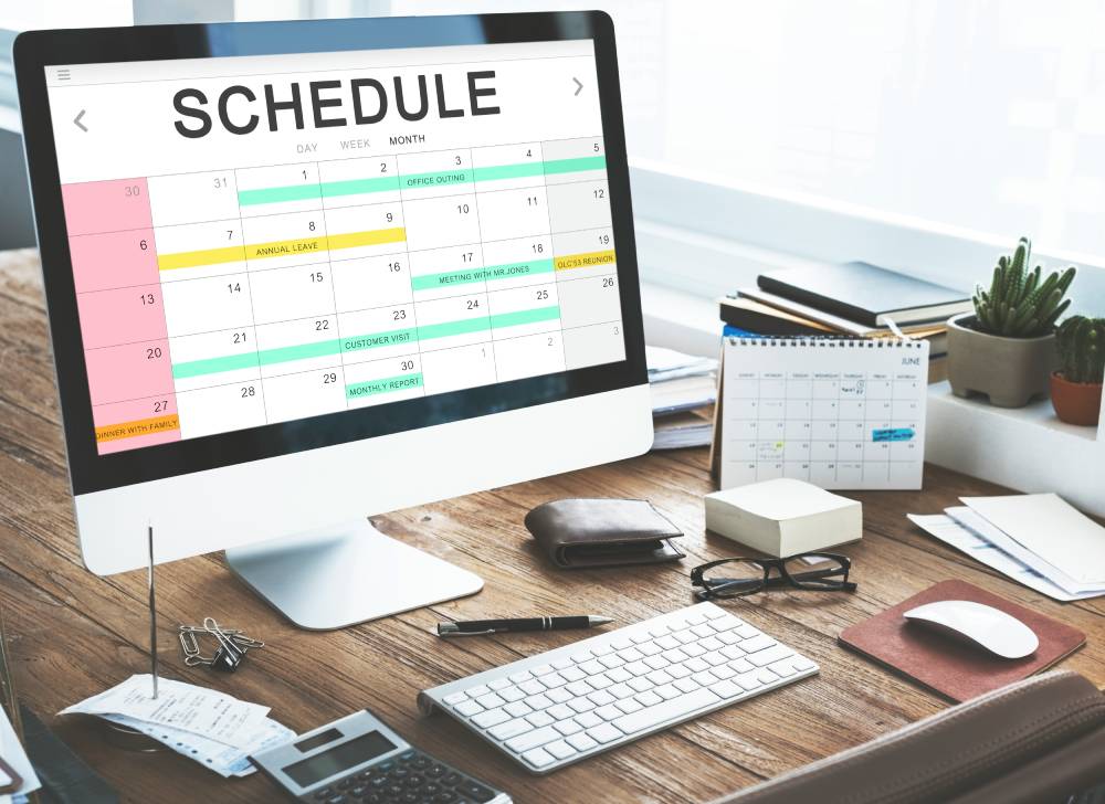 A computer monitor displaying a calendar schedule sits on a wooden desk alongside a keyboard, mouse, glasses, and papers. The setup creates an organized workspace for those seeking services from a notary near Kissimmee, FL.