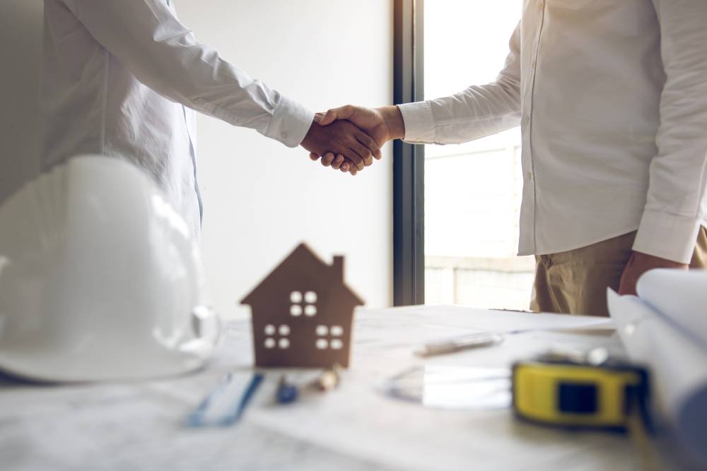 Two people in white shirts shake hands over a desk featuring a small house model, hard hat, and construction plans, symbolizing an agreement or partnership in a construction project. This meeting conveniently takes place near Kissimmee with the assistance of a local notary to finalize documents.