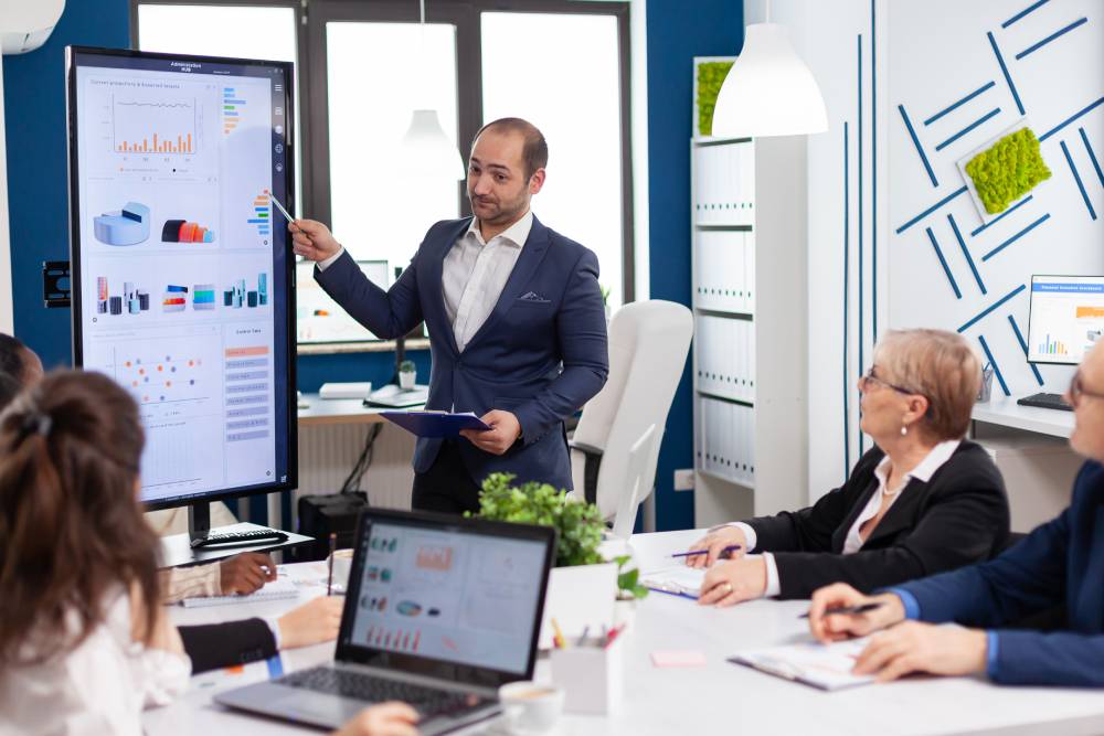 A man in a suit, possibly a notary near Kissimmee, presents charts on a screen to a focused group seated at a conference table filled with laptops and documents.