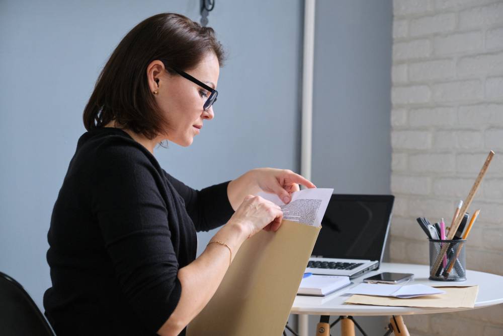 A person wearing glasses, focused on tax preparation near Kissimmee, places a document into a folder at a desk equipped with a laptop, notebook, pencil holder, and smartphone.