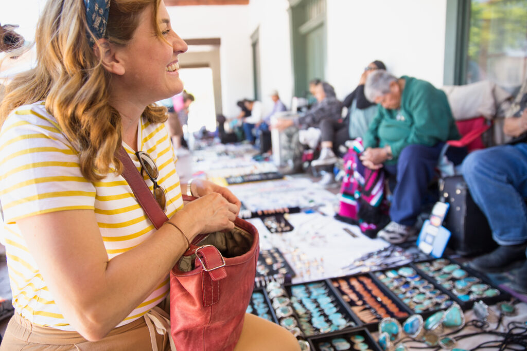 Woman in striped shirt smiles while looking at jewelry displayed on a blanket at an outdoor market.