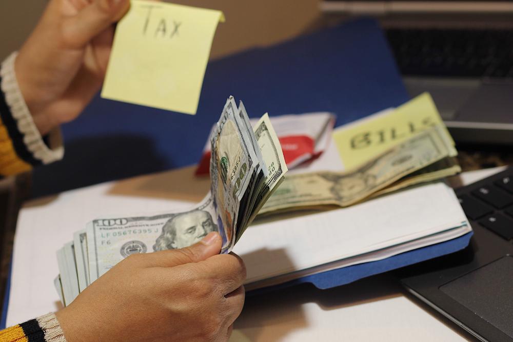 A person holding dollar bills with sticky notes labeled "TAX" and "BILLS" near a laptop, perhaps getting ready for tax preparation near Kissimmee, FL.