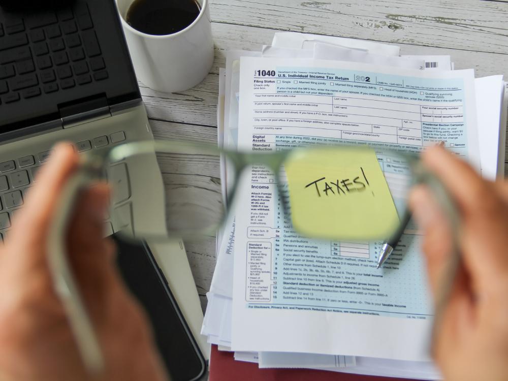 Hands hold glasses focusing on a sticky note labeled "TAXES!" over a stack of IRS 1040 forms near a laptop and a cup of coffee, as the urgency of tax preparation near Kissimmee, FL, sets in.